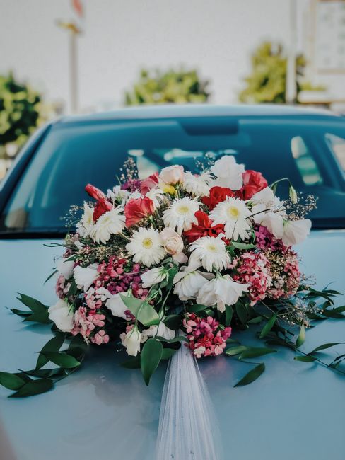 A large, vibrant floral bouquet placed on the hood of a light blue car, featuring a mix of white daisies, pink and red roses, white lilies, and small clusters of pink and purple flowers, all accented 