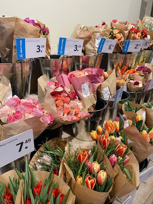 A display of various floral bouquets arranged in metallic buckets and wrapped in brown craft paper, showcasing a range of vibrant flowers including pink roses, peach and coral carnations, orange and p