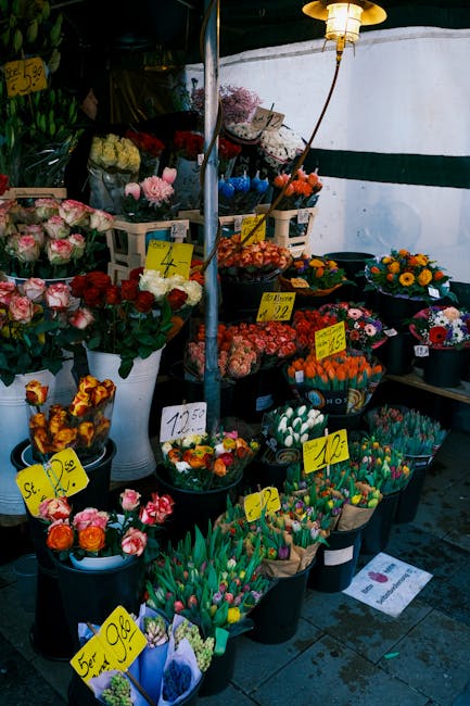 A vibrant outdoor flower stall featuring an assortment of fresh floral bouquets and arrangements. The display includes pink and white roses, multicolored tulips, orange and yellow marigolds, and vario