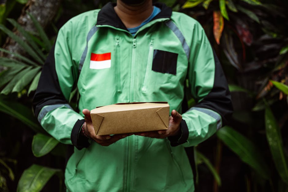A person wearing a green delivery jacket with a red and white flag patch and black accents, holding a rectangular cardboard box with folded corners, standing outdoors amidst lush green foliage. The in
