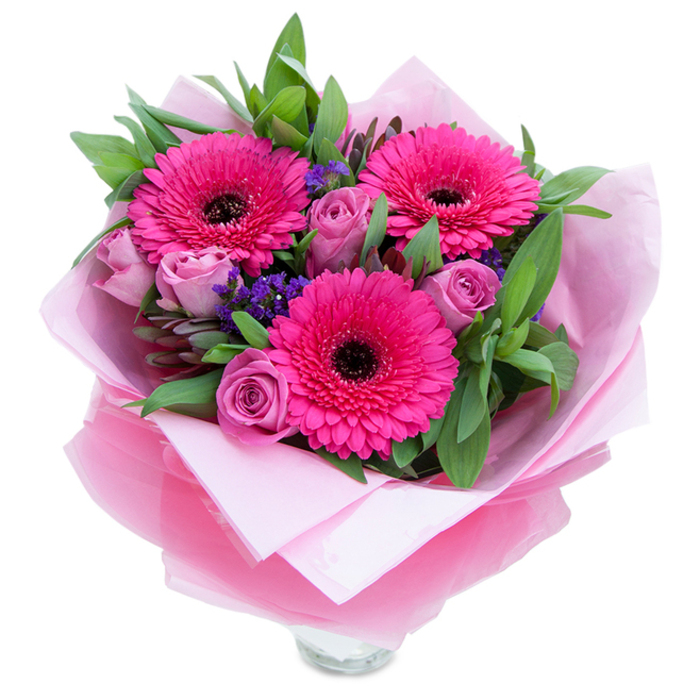 I always respond to pinks that balance playfulness with quiet grace, and this bouquet does exactly that. Three wide-faced Gerbera daisies in a saturated fuchsia burst open at the heart, their many petals forming a confident halo around each dark centre. Nestled close are roses in a gentle lavender-pink or dusty rose, their petals still tender and curled, suggesting both newness and intimacy. Small sprays of purple statice weave through the blooms like little notes of contrast, adding texture and a wildflower spirit against broad, verdant foliage that reads like salal or eucalyptus-leaves with a cool, slightly waxy sheen that hold light and hint at a fresh, garden aroma. The flowers are swaddled in a double layer of wrap: a soft, matte pale pink tissue that feels almost like fabric beneath the stems, and an outer layer of sheer, crinkled fuchsia-pink cellophane that refracts light and ties the pink palette together. At the base, the top rim of a clear glass vase is exposed, a small promise that the arrangement is meant to sit and be admired in a living room or on a dining table. The overall effect is intimate and uplifting, evoking the quiet beauty of a walk through Ruislip Woods at dawn-gentle, composed, and capable of brightening a bedside or serving as a heartfelt gift for someone special. Every fold and stem placement reflects a commitment to quality and the kind of careful making local customers appreciate.
